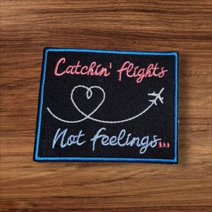 May include: A black square patch with a blue border and the text "Catchin' flights Not feelings..." in pink and white script. A white heart and airplane graphic are also featured.