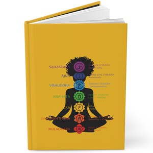 May include: A yellow journal with a black silhouette of a person in a yoga pose. The person has seven chakras represented by colored circles with Sanskrit symbols. The chakras are labeled with their names and descriptions.