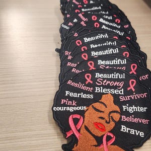 May include: Black and pink embroidered patch with a woman's face and the words "Beautiful Strong Blessed Survivor Fighter Believer Brave" and "Resilient Fearless Pink Courageous".