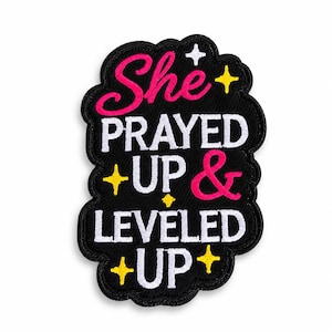 May include: A black embroidered patch with a scalloped edge featuring the pink text She, white text PRAYED, pink ampersand, and white text UP & LEVELED UP. The design is accented with small yellow and white four-pointed star icons throughout.