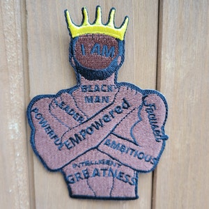 May include: A brown embroidered patch depicting a person with a crown on their head and the words "I AM BLACK MAN LEADER EMPOWERED INTELLIGENT POWERFUL AMBITIOUS  EQUAL  GREATNESS" around them.
