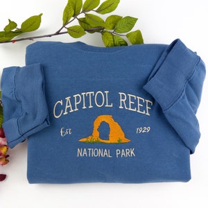 Puede incluir: Una sudadera azul con el texto "CAPITOL REEF NATIONAL PARK" y un gráfico de arco naranja. El texto "Est 1929" también está incluido.