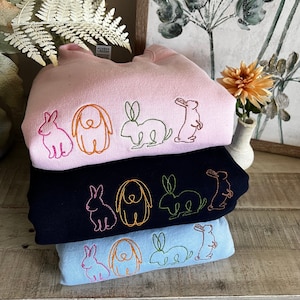 May include: A stack of three sweatshirts with embroidered images of rabbits and dogs. The top sweatshirt is pink, the middle sweatshirt is black, and the bottom sweatshirt is light blue.