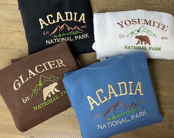 May include: Four crewneck sweatshirts in black, white, brown, and blue. Each features embroidered text and graphics of national parks including Acadia, Yosemite, and Glacier, with mountain silhouettes, bear figures, and established dates.