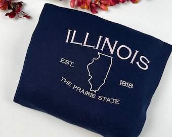 May include: A navy blue sweatshirt with a white embroidered design featuring the state of Illinois, the text "ILLINOIS", "EST. 1818", and "THE PRAIRIE STATE".