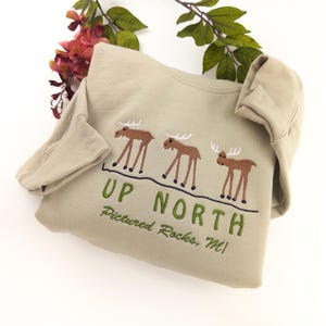 May include: A tan sweatshirt with a green embroidered design of three moose and the text "UP NORTH Pictured Rocks, MI".