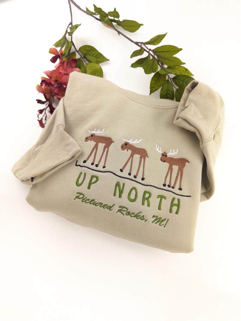 May include: A tan sweatshirt with a green embroidered design of three moose and the text "UP NORTH Pictured Rocks, MI".