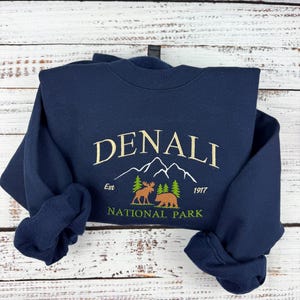 Op de afbeelding: Een marineblauwe sweatshirt met het woord "DENALI" in goud geborduurd, samen met een bergketenafbeelding, een eland, een beer en de tekst "NATIONAL PARK". De sweatshirt heeft ook de tekst "Est 1917".