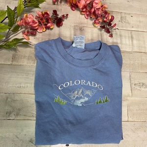 K&ouml;nnte beinhalten: Ein hellblaues T-Shirt mit einem gestickten Design aus Bergen und B&auml;umen. Der Text "COLORADO" ist &uuml;ber dem Design gestickt.
