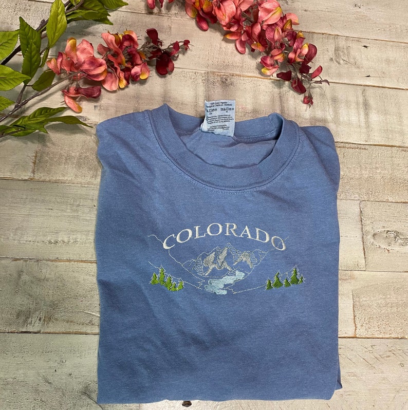 K&ouml;nnte beinhalten: Ein hellblaues T-Shirt mit einem gestickten Design aus Bergen und B&auml;umen. Der Text "COLORADO" ist &uuml;ber dem Design gestickt.