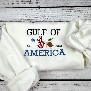 A white sweatshirt with the text 'GULF OF AMERICA' embroidered in black and blue. The text is surrounded by small embroidered images of a coral, a fish, and a sea turtle. The text also includes the words 'Est' and '2025'.