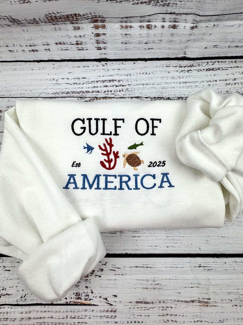 A white sweatshirt with the text 'GULF OF AMERICA' embroidered in black and blue. The text is surrounded by small embroidered images of a coral, a fish, and a sea turtle. The text also includes the words 'Est' and '2025'.