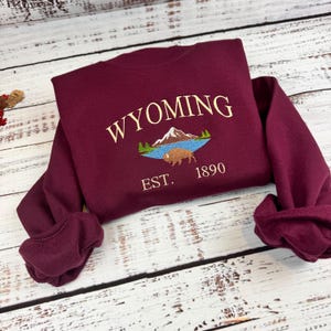 May include: A maroon long-sleeve sweatshirt featuring an embroidered design of a mountain range, a blue lake, and a brown bison. The text "WYOMING" is arched above the graphic, and "EST. 1890" is embroidered in gold thread below the bison.