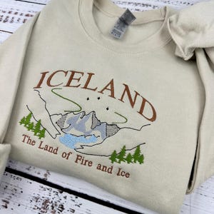 Puede incluir: Una sudadera beige con un dise&ntilde;o bordado de una cordillera y el texto "ICELAND The Land of Fire and Ice".