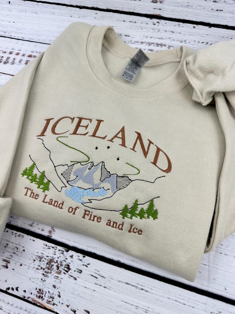 Puede incluir: Una sudadera beige con un dise&ntilde;o bordado de una cordillera y el texto "ICELAND The Land of Fire and Ice".