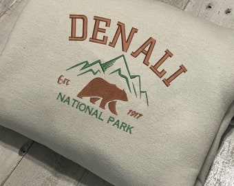 May include: A beige sweatshirt with an embroidered logo for Denali National Park. The logo features a brown bear, green mountains, and the text "Denali National Park Est. 1917".