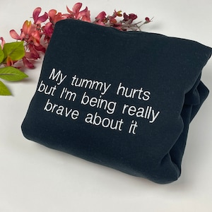 Puede incluir: Una sudadera negra doblada con texto bordado en blanco que dice "My tummy hurts but I'm being really brave about it". La sudadera se muestra sobre una superficie blanca con acentos florales rosas y verdes.