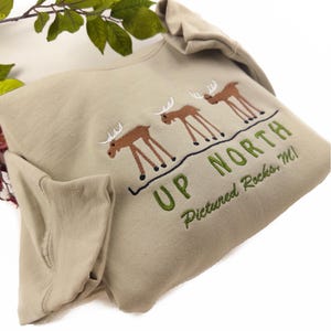 May include: A tan long-sleeve shirt with a green and brown embroidered design of three moose and the text "Up North Pictured Rocks MI".