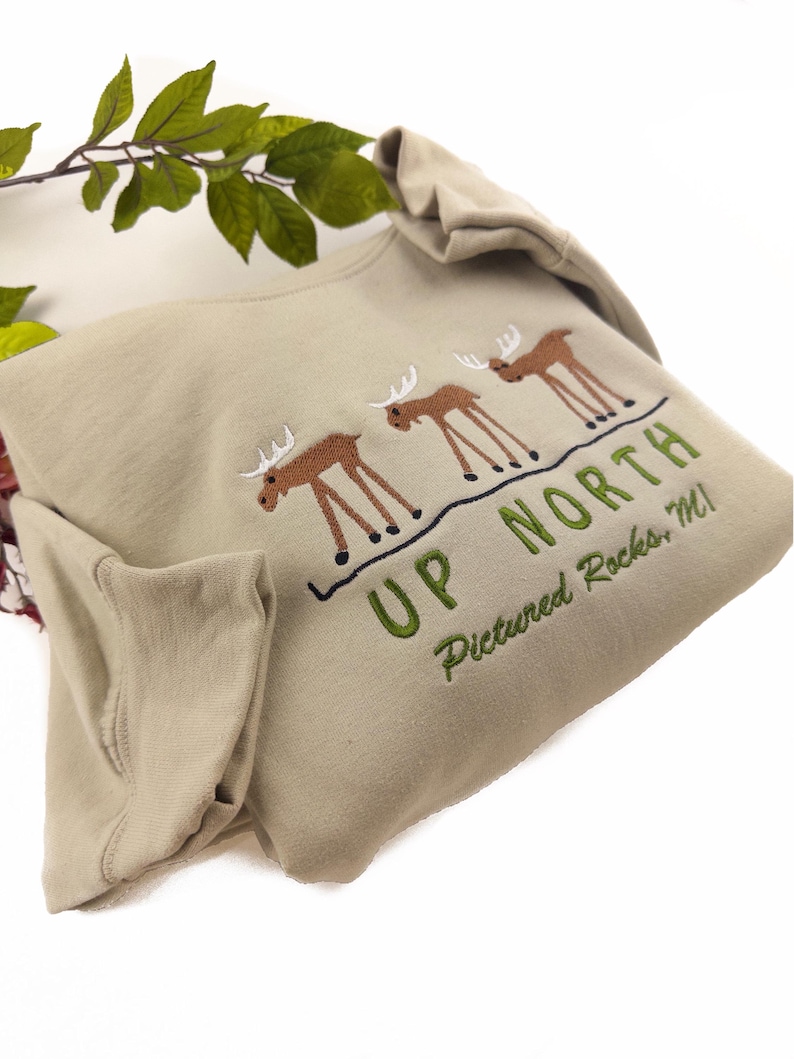 May include: A tan long-sleeve shirt with a green and brown embroidered design of three moose and the text "Up North Pictured Rocks MI".