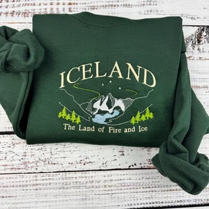 Puede incluir: Una sudadera verde oscuro con un dise&ntilde;o bordado en blanco que dice "ICELAND The Land of Fire and Ice".