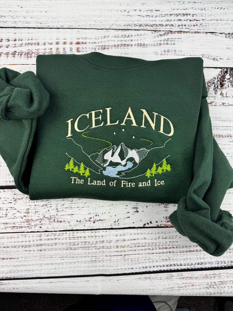 Puede incluir: Una sudadera verde oscuro con un dise&ntilde;o bordado en blanco que dice "ICELAND The Land of Fire and Ice".