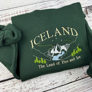 Puede incluir: Una sudadera verde oscuro con un dise&ntilde;o bordado de una cordillera y el texto "ICELAND The Land of Fire and Ice".