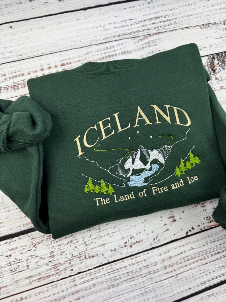 Puede incluir: Una sudadera verde oscuro con un dise&ntilde;o bordado de una cordillera y el texto "ICELAND The Land of Fire and Ice".