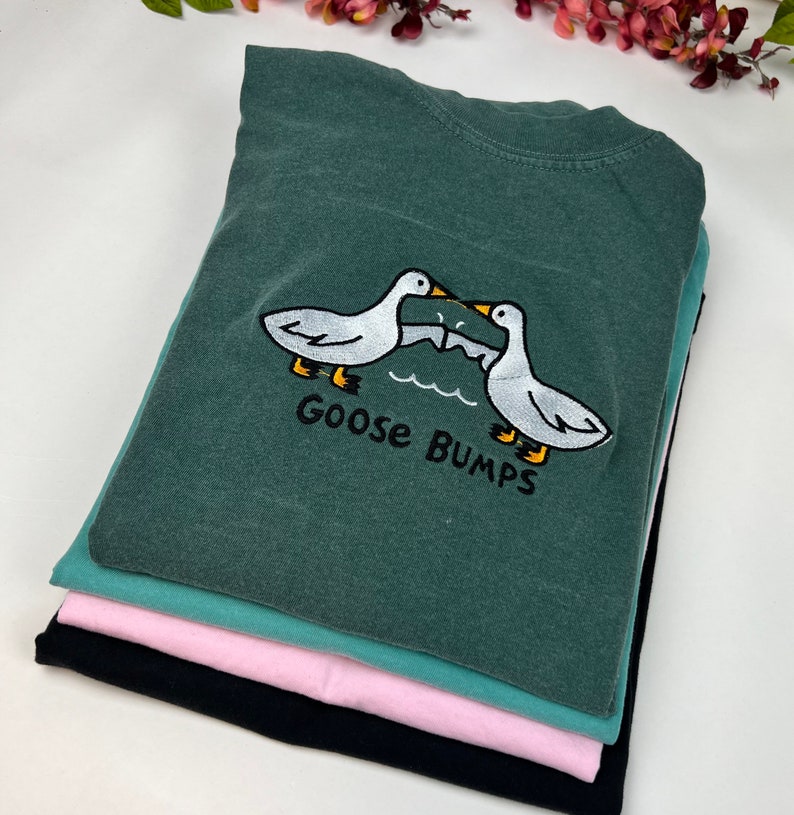 May include: A stack of four t-shirts, the top one is dark green with a white embroidered design of two geese facing each other with the text "Goose Bumps" underneath.