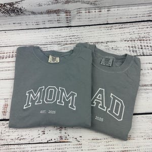 Może przedstawiać: Dwie szare koszulki z napisami "MOM" i "AD" w kolorze białym, z nadrukowanym rokiem "2025". Koszulka z napisem "MOM" ma nadrukowane słowa "EST. 2025".