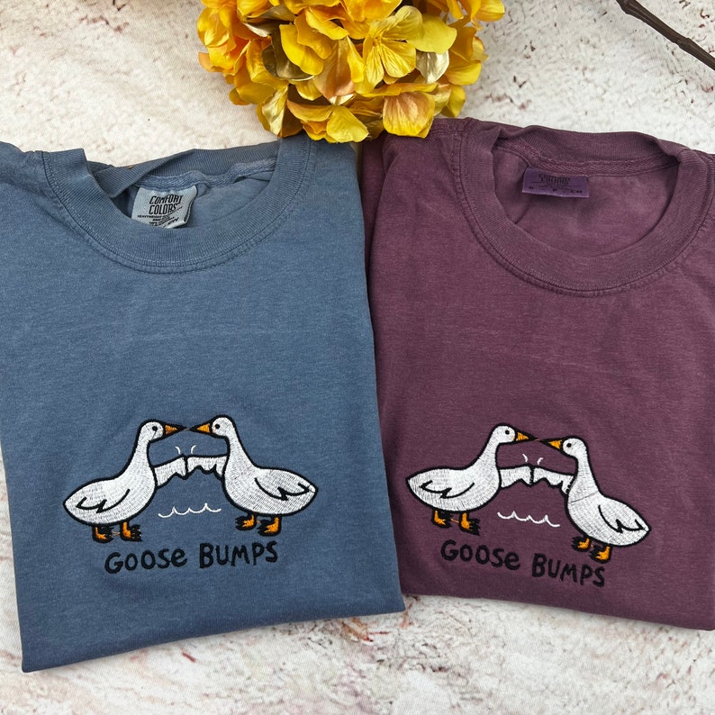 May include: Two t-shirts with a goose bump design. The shirts are in blue and purple. The design features two geese with their wings outstretched, facing each other. The text "Goose Bumps" is printed below the geese.