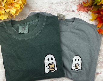 May include: Two t-shirts with a ghost design. The ghost on the left is holding a cup of coffee with the word "COFFEE" printed on it. The ghost on the right is holding a cup with a straw. The shirt on the left is a dark green color and the shirt on the right is a light gray color.