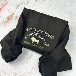 May include: Black sweatshirt with a white embroidered design of a lion in front of mountains. The text "NGORONGORO CONSERVATION AREA Est. 1959" is embroidered in white.