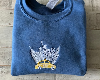 May include: A blue sweatshirt with a white embroidered design of the New York City skyline and a yellow taxi cab with the letters "NYC" on the side.