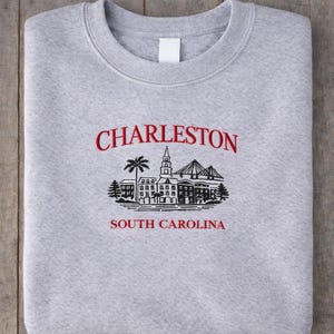 以下が含まれることがあります： 折りたたまれたライトグレーのスウェットシャツ。赤い文字で「CHARLESTON」と書かれ、その下にチャールストンのランドマークの白黒イラストが描かれています。イラストの下には「SOUTH CAROLINA」の文字も赤色で書かれています。