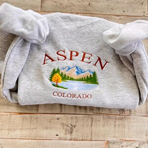 May include: A heather gray sweatshirt with the embroidered word "ASPEN" in maroon above a scenic mountain and lake design. The word "COLORADO" is embroidered below the design. The sweatshirt has a crew neck and long sleeves.