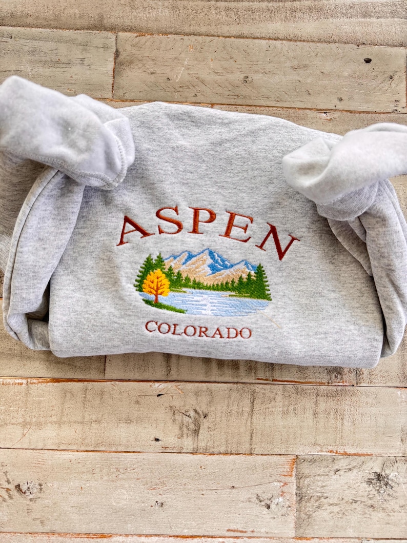 May include: A heather gray sweatshirt with the embroidered word "ASPEN" in maroon above a scenic mountain and lake design. The word "COLORADO" is embroidered below the design. The sweatshirt has a crew neck and long sleeves.