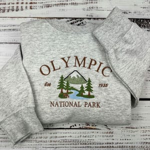 Puede incluir: Una sudadera gris con la inscripción bordada "OLYMPIC NATIONAL PARK" en marrón y verde. El diseño incluye una montaña, árboles y un río. La sudadera también tiene bordado el año "1935".
