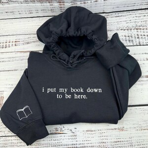 Op de afbeelding: Zwart hoodie met de tekst "i put my book down to be here." in witte letters. De linkermouw heeft een klein geborduurd beeld van een open boek.