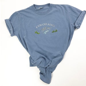 A blue t-shirt with a white embroidered design of mountains and trees. The text 'COLORADO' is embroidered in white above the mountains.