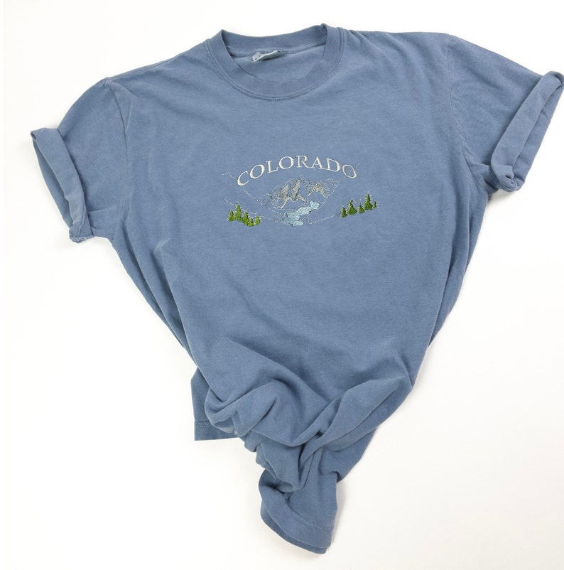A blue t-shirt with a white embroidered design of mountains and trees. The text 'COLORADO' is embroidered in white above the mountains.