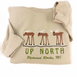 May include: A beige long-sleeved sweatshirt with a green embroidered design of three moose and the text "UP NORTH Pictured Rocks, MI".