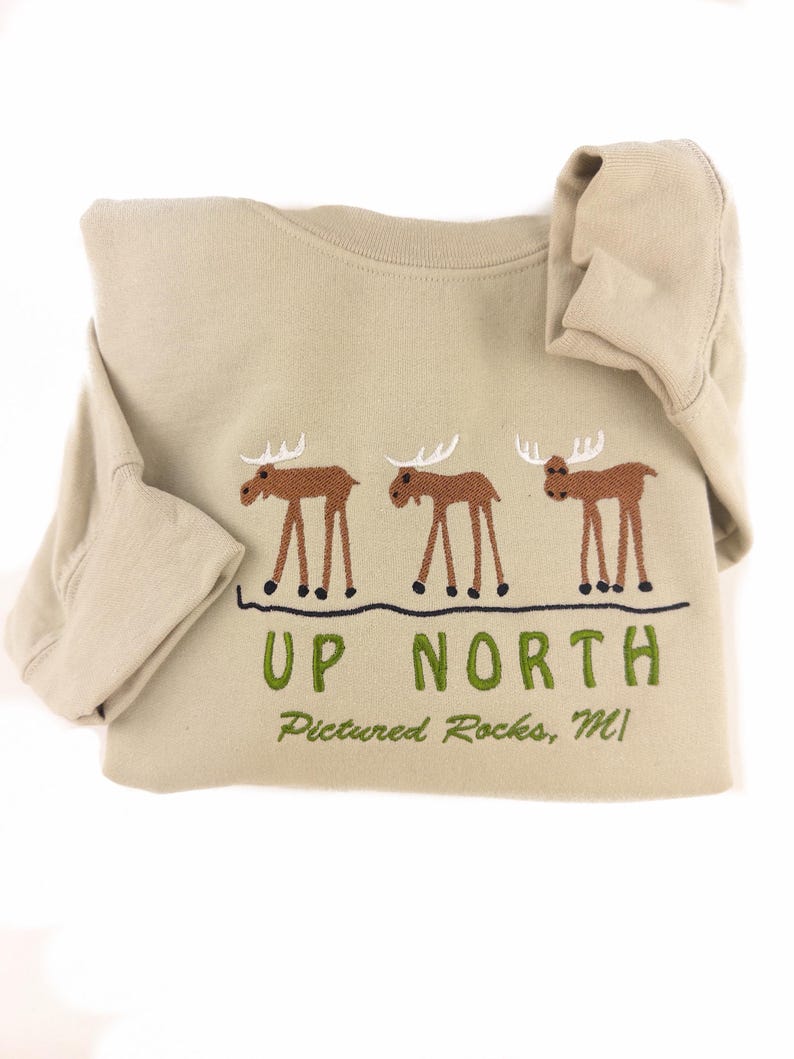 May include: A beige long-sleeved sweatshirt with a green embroidered design of three moose and the text "UP NORTH Pictured Rocks, MI".