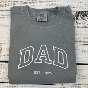 Może przedstawiać: Szary T-shirt z napisem "DAD" w białych, obwiedzionych literach i tekstem "EST. 2025" poniżej. Koszulka jest marki Comfort Colors.