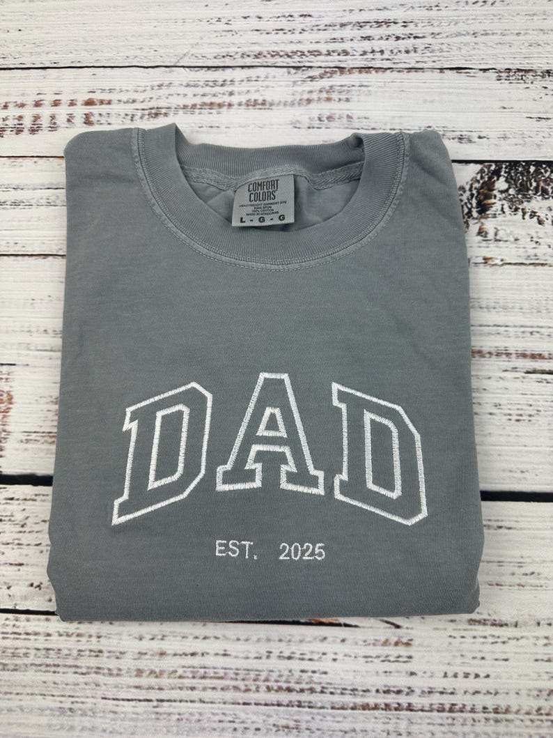 Może przedstawiać: Szary T-shirt z napisem "DAD" w białych, obwiedzionych literach i tekstem "EST. 2025" poniżej. Koszulka jest marki Comfort Colors.