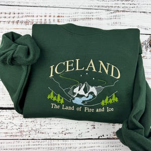 Puede incluir: Una sudadera verde oscuro con la palabra "ICELAND" bordada en color amarillo claro. La sudadera tambi&eacute;n presenta una imagen bordada de una cordillera con un lago y el texto "The Land of Fire and Ice".