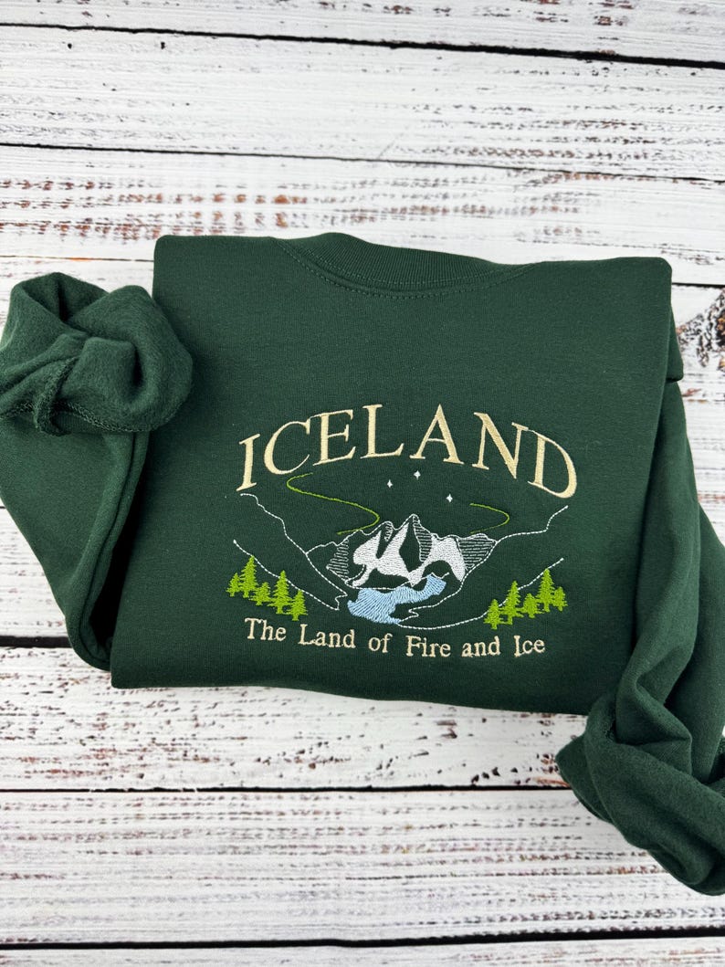 Puede incluir: Una sudadera verde oscuro con la palabra "ICELAND" bordada en color amarillo claro. La sudadera tambi&eacute;n presenta una imagen bordada de una cordillera con un lago y el texto "The Land of Fire and Ice".