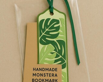 May include: A handmade monstera bookmark with a green leaf design. The bookmark is made of layered paper and has a green ribbon attached. The bookmark is packaged in a clear plastic sleeve with a brown tag that reads "HANDMADE MONSTERA BOOKMARK."