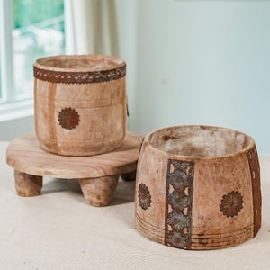 May include: Two wooden planters with decorative metal accents. The larger planter has a floral design and the smaller planter has a circular design. Both planters are sitting on a wooden stand with four legs.