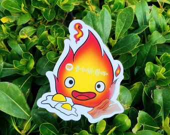 May include: A sticker featuring a cartoon fire spirit with a smiling face, holding a plate of bacon and eggs. The fire spirit is red and orange with a yellow face and black eyes. The sticker has a white background and a Spotify code.