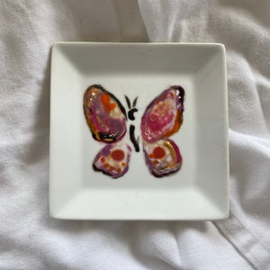 May include: Square white ceramic dish with a hand-painted butterfly design. The butterfly features purple, pink, orange, and gold accents. The dish is a small size, perfect for holding jewelry or small items.